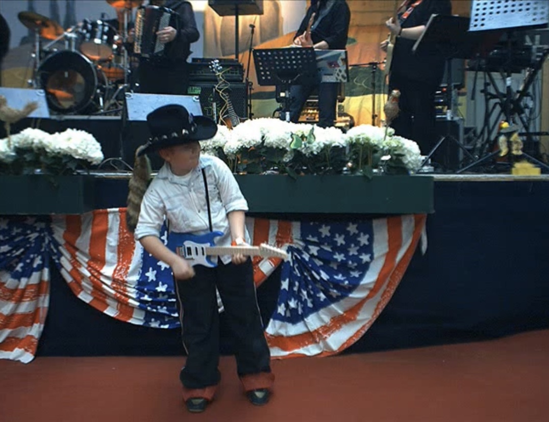 Child dressed in white shirt, black hat, and black pants holding a toy guitar on stage with musical instruments and American flag-themed decorations behind.