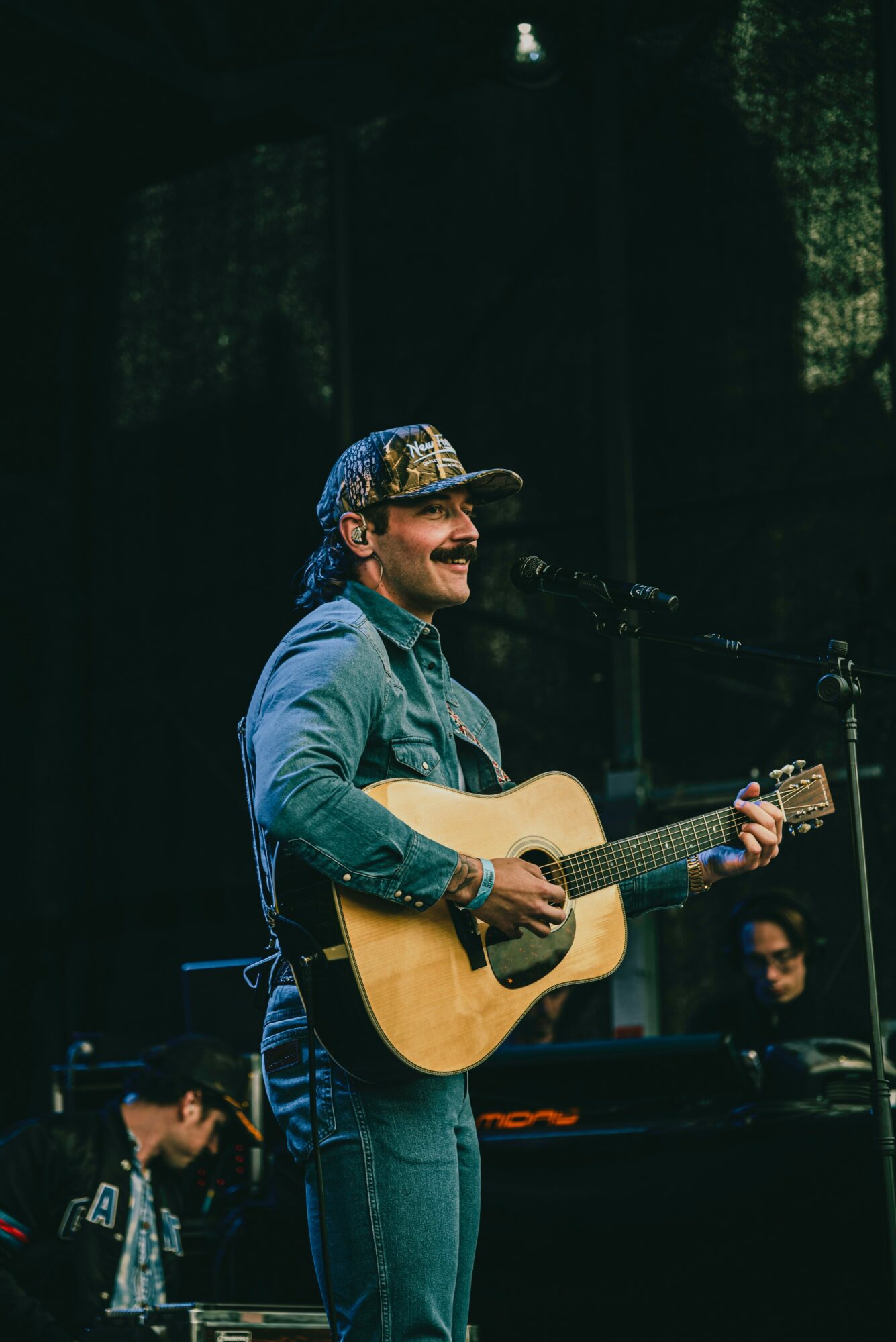 Person playing guitar and singing on stage with a microphone, wearing a cap and denim shirt.