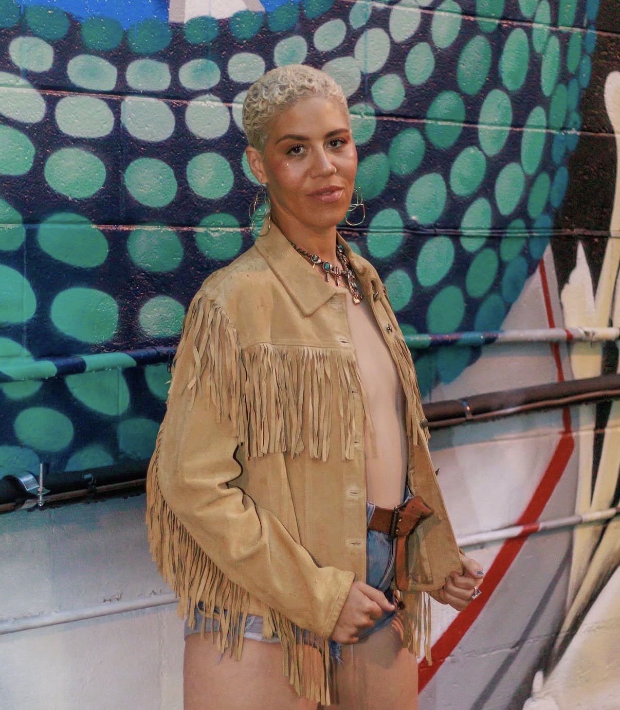 Woman with short, platinum blonde hair wearing a fringed tan jacket and jeans, standing in front of a colorful mural.