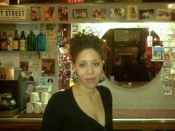 Young woman with curly hair and earrings in a store with shelves of bottles and photos behind her.