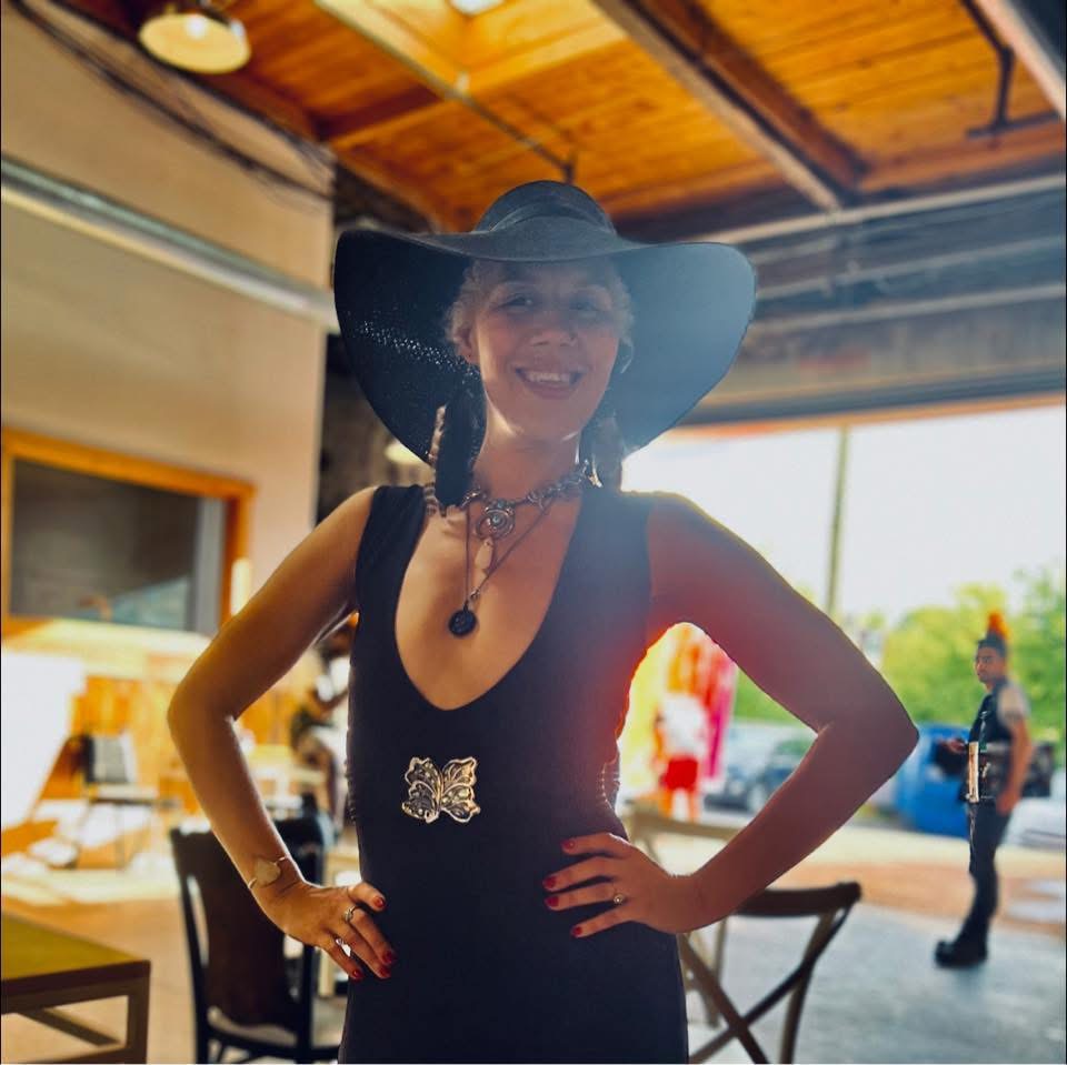 Woman wearing a large hat and black dress, smiling indoors with outdoor view in background.