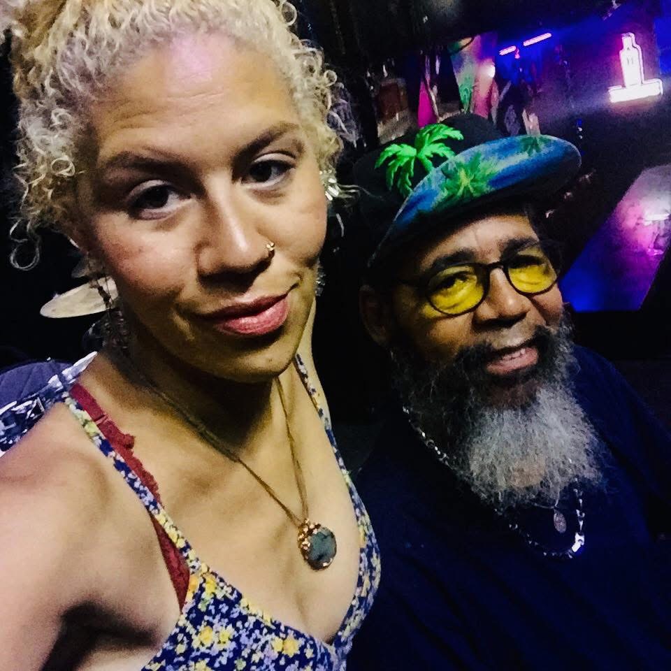 Two people taking a selfie in a dimly lit, colorful environment, one with curly blonde hair and the other with a hat and glasses.
