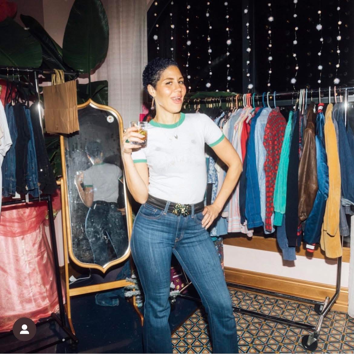 Woman holding a drink, standing in a room with clothing racks and a mirror, smiling, casual outfit, indoor setting.