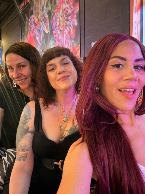 Three women posing together indoors, smiling, with colorful background and floral decor.