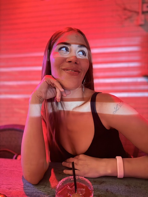 Woman with long hair smiling, resting her chin on her hand, in a dimly lit setting with red lighting, wearing a black top.