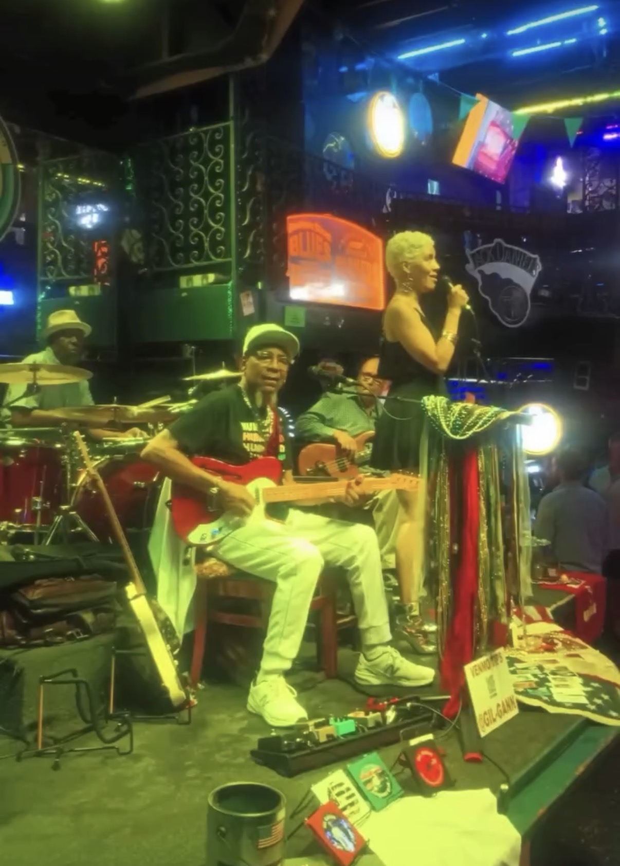 Three musicians perform on stage with a woman singing, colorful lights, and electronic equipment in the background.