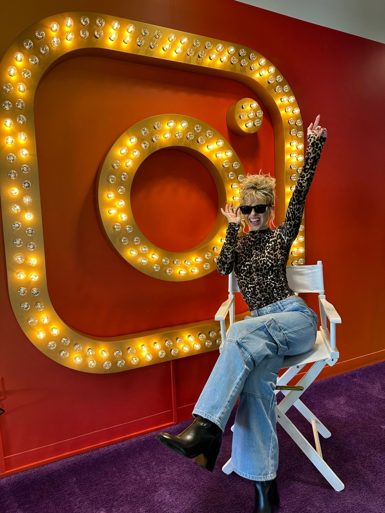 Person lying on a large illuminated Instagram logo with a red background and purple carpet, wearing a patterned top and jeans.
