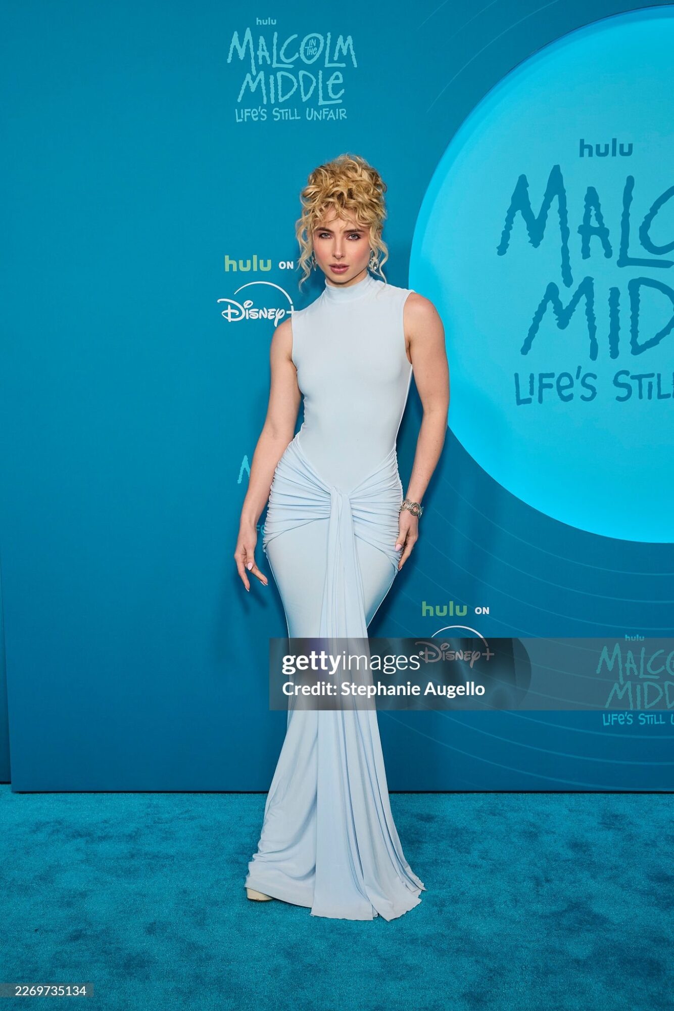 Woman in white dress standing on blue carpet at event backdrop with text, facing forward.