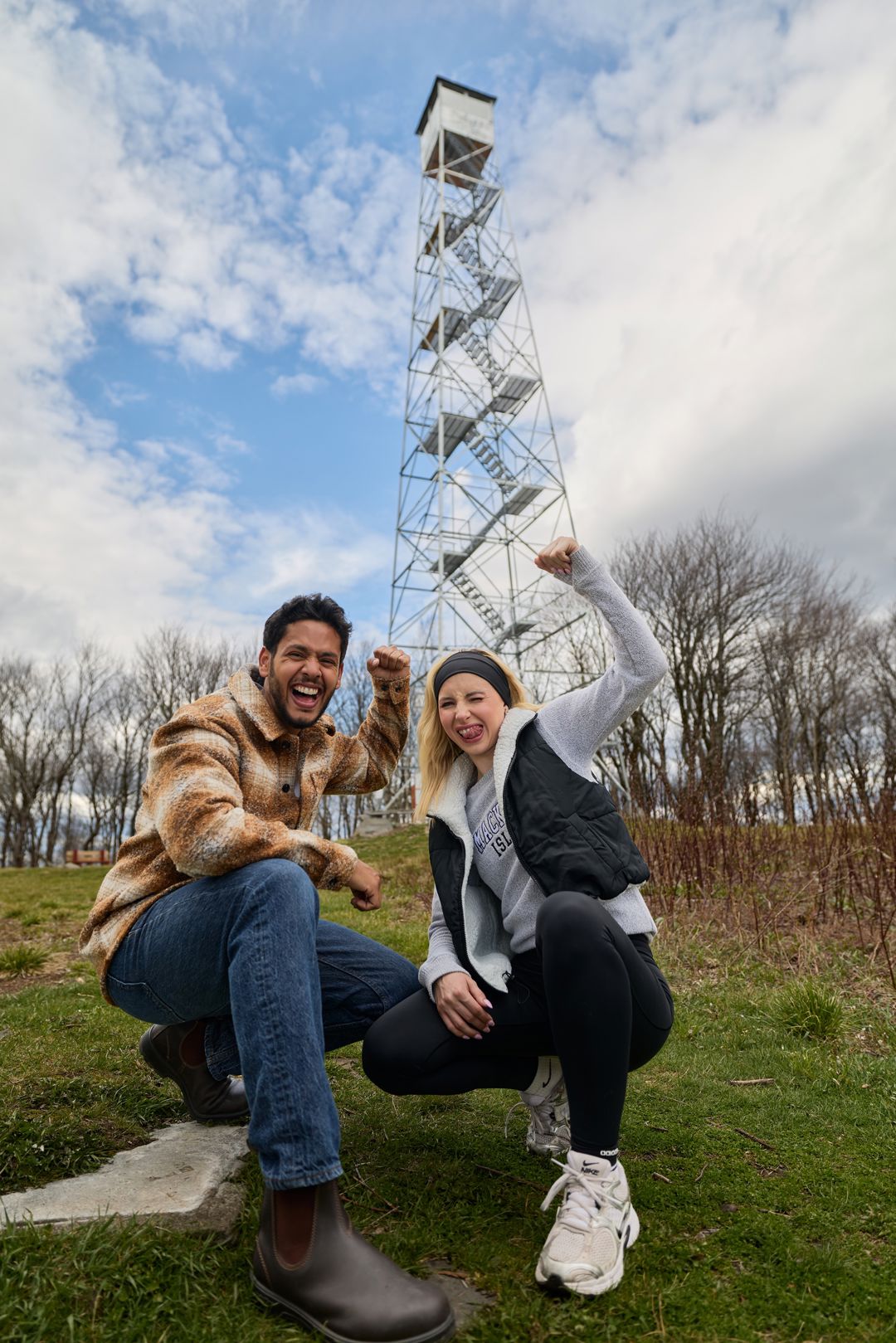 Two people smiling and celebrating outdoors near a tall metal tower, with trees and a partly cloudy sky in the background.