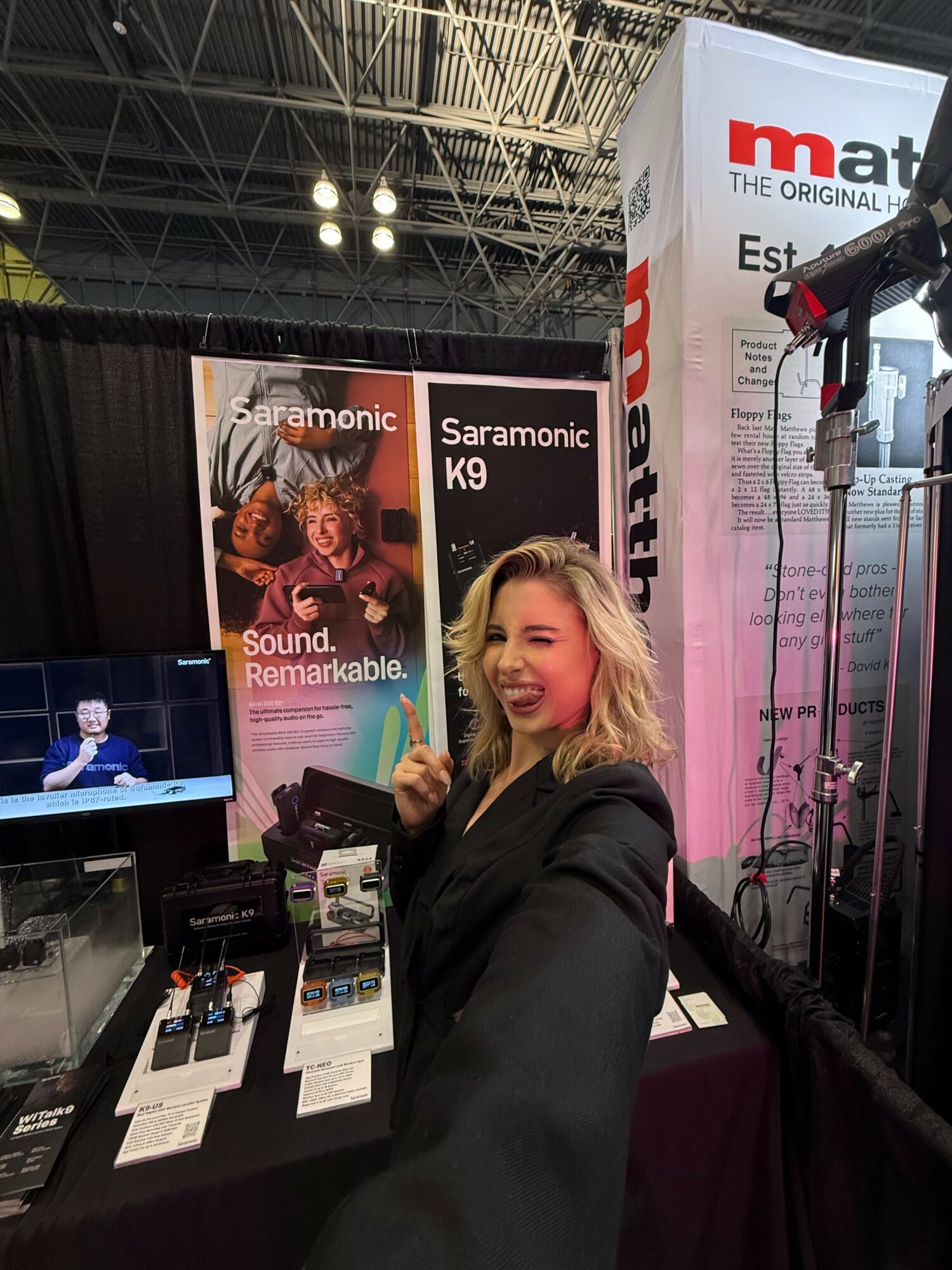 Woman smiling and giving a thumbs-up at a trade show booth with Saramonic K9 products, banners, and equipment in the background.