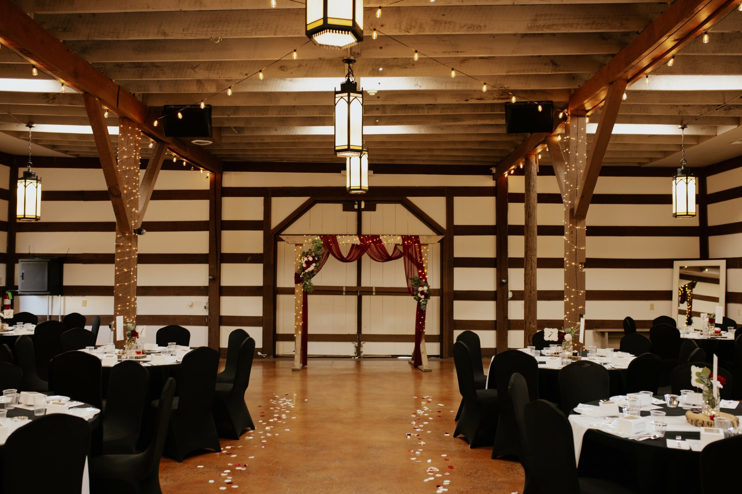 Event space with decorated arch, tables with black chairs, and hanging lights, ready for a celebration or wedding reception.
