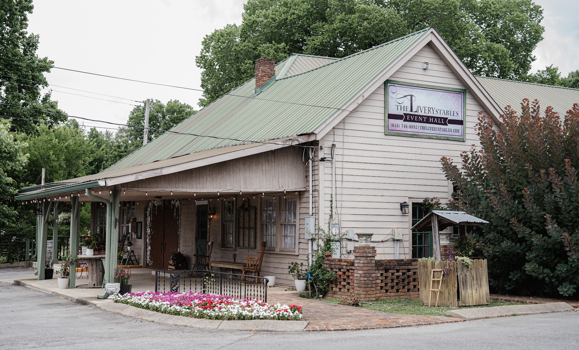 Old building with a green roof, porch, and a sign, surrounded by trees and a flower bed.