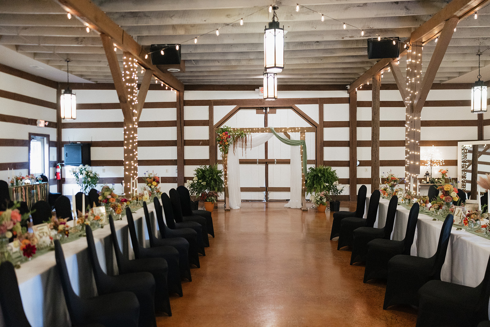 Wedding reception hall with long tables, black chairs, floral arrangements, and wooden beams with string lights.