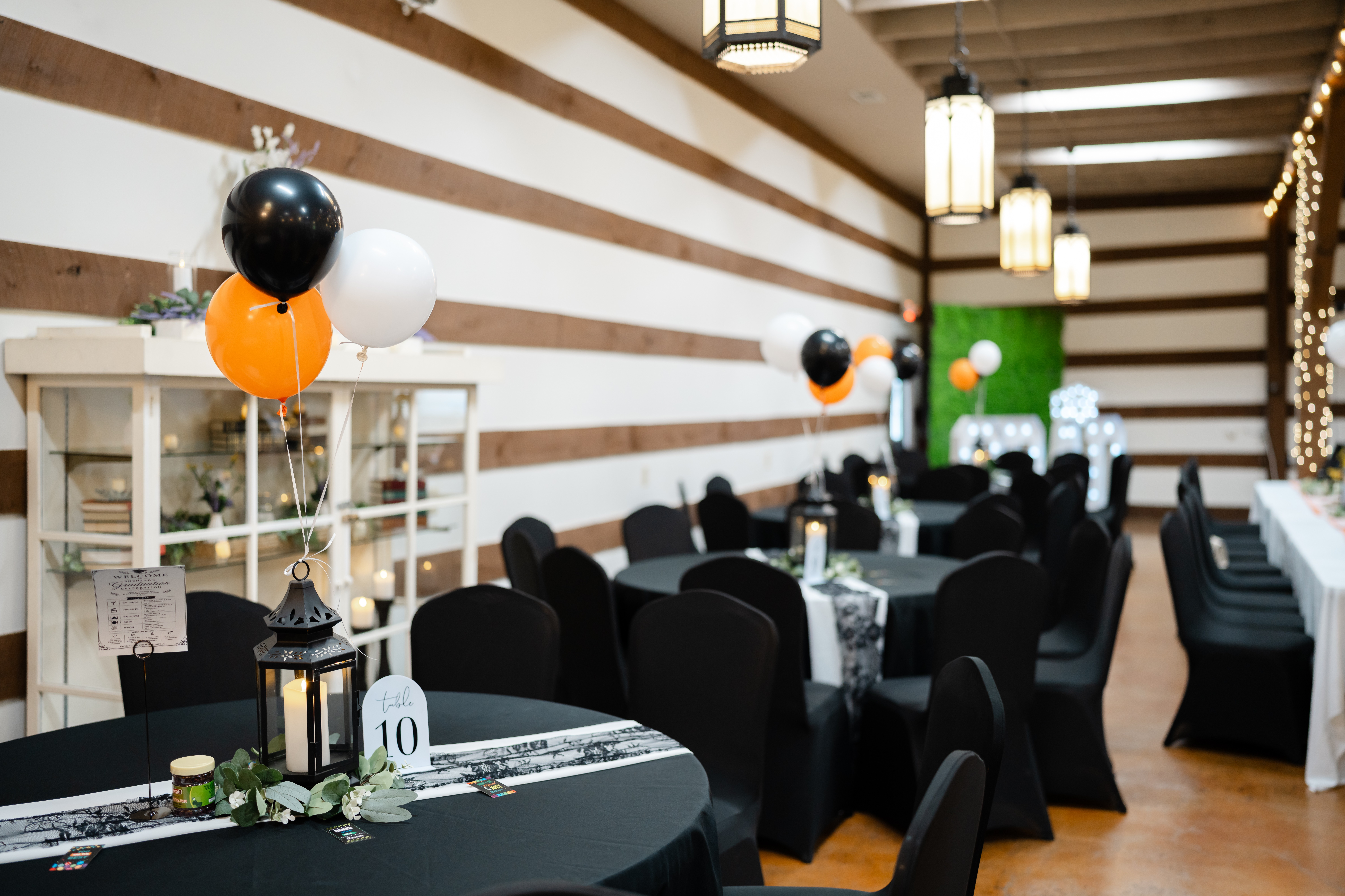 Party room with black chairs, decorated tables, balloons, and string lights, featuring a wooden wall and hanging lanterns.