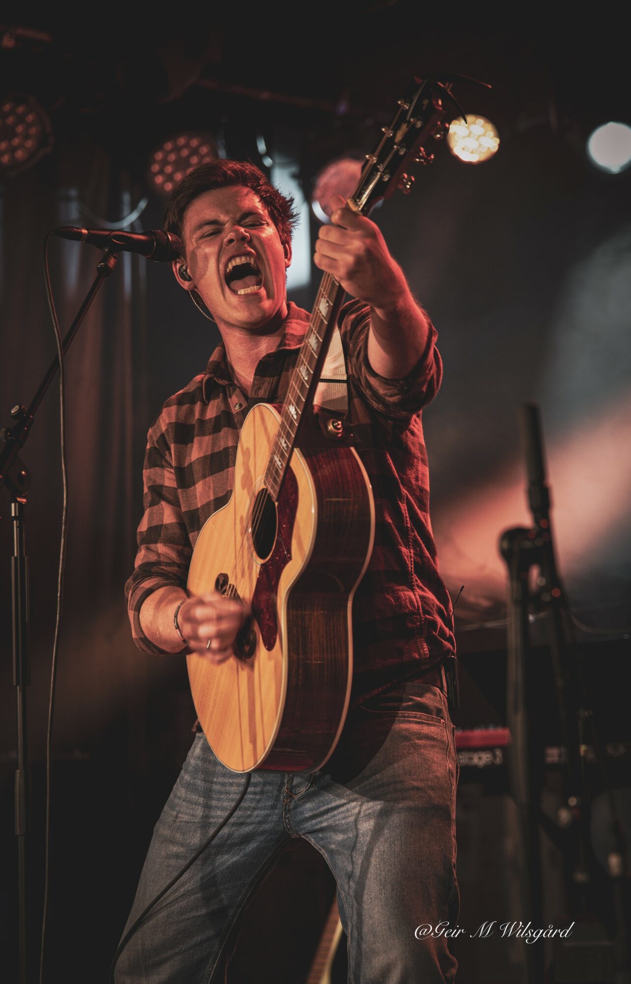 Person passionately singing and playing guitar on stage with stage lights in background