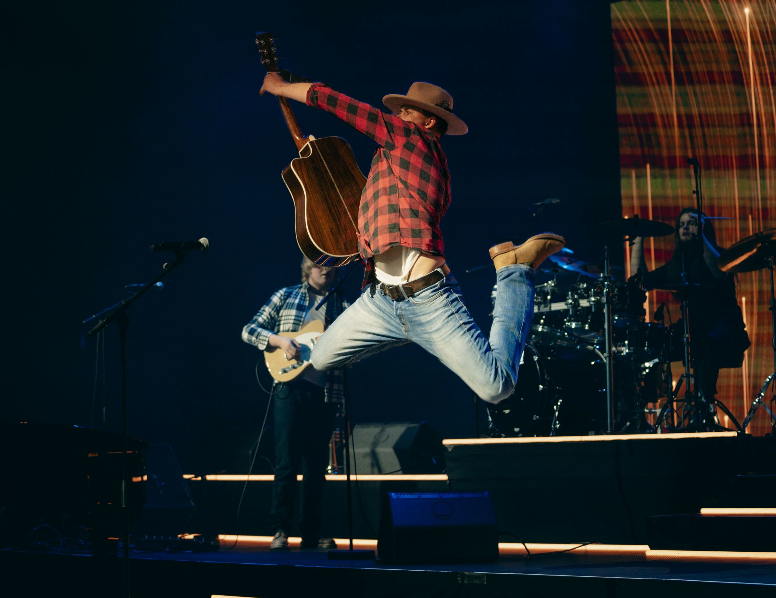 Person jumping on stage with guitar, wearing a hat and plaid shirt, performing with band members in background.