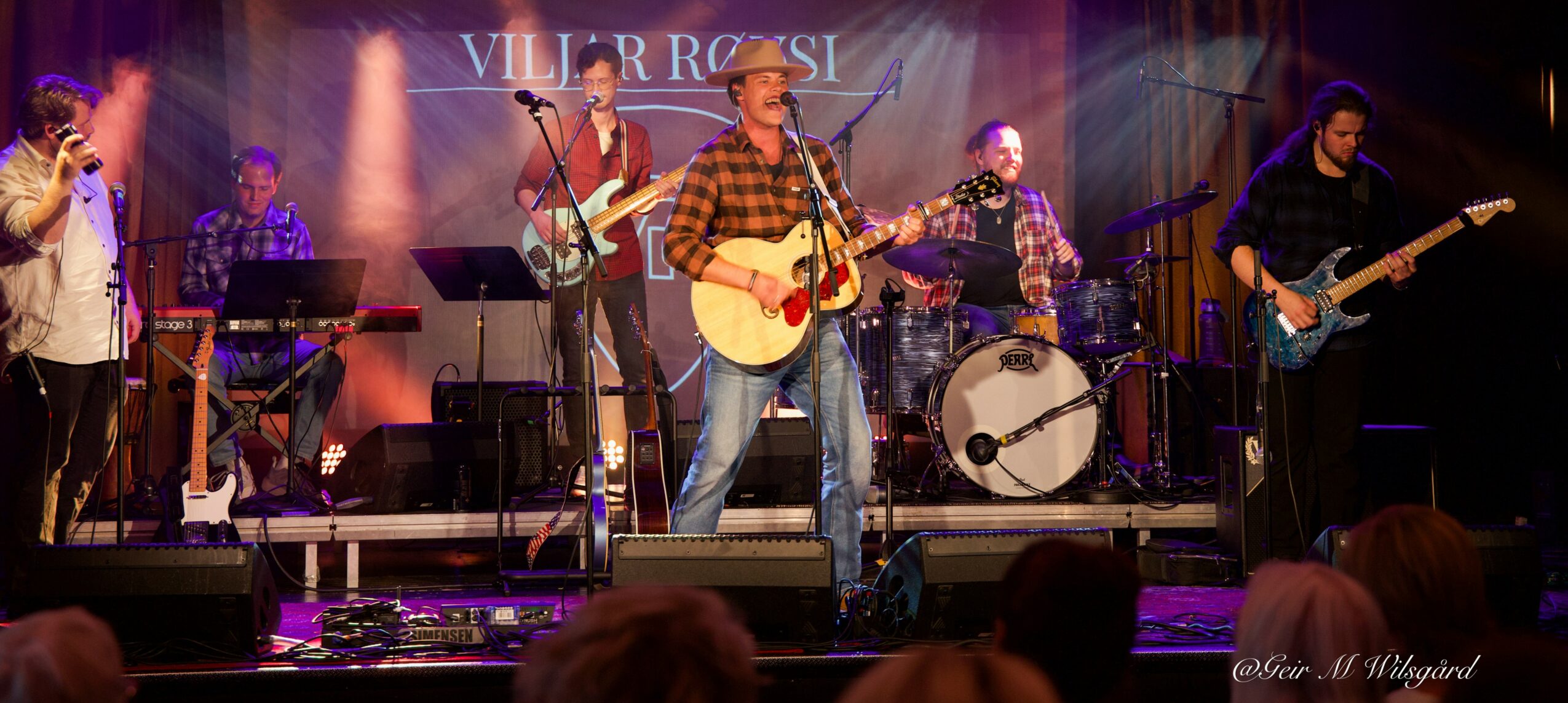 Band performs on stage with five musicians, one singing and playing guitar, others playing keyboard, drums, and bass, with stage lighting.