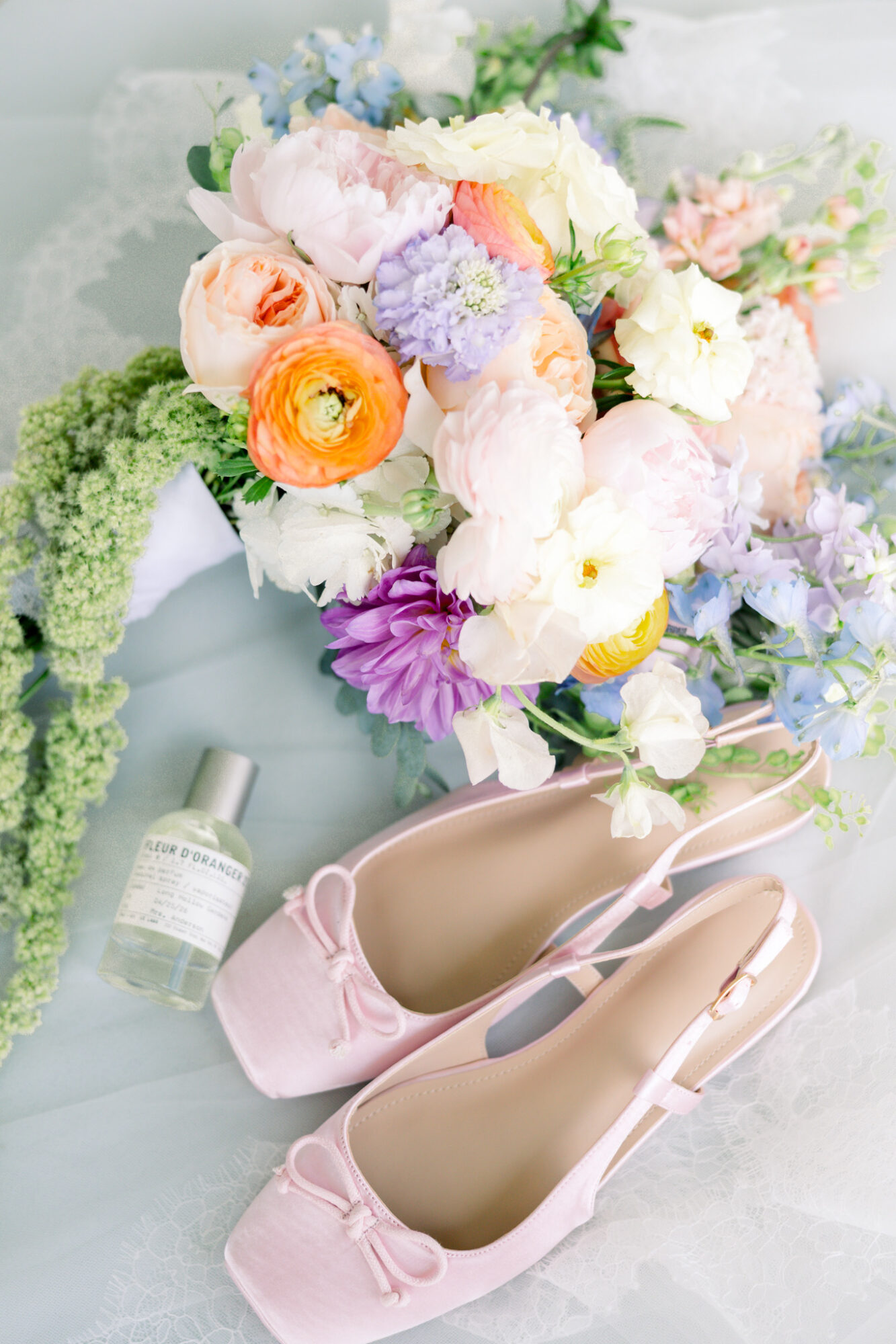 Pink high-heeled shoes with a bow, a bouquet of pastel flowers, and a small bottle on a light surface.