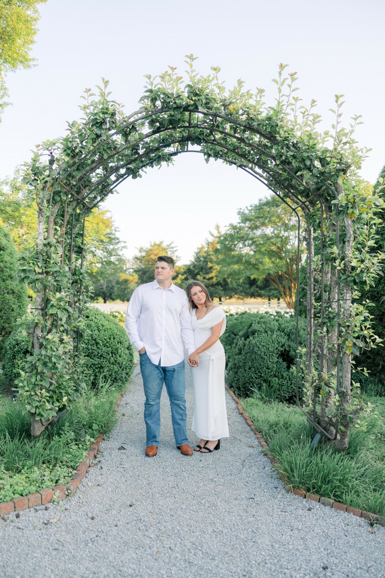 A couple stands under a green archway in a park, surrounded by trees and bushes, on a gravel path.