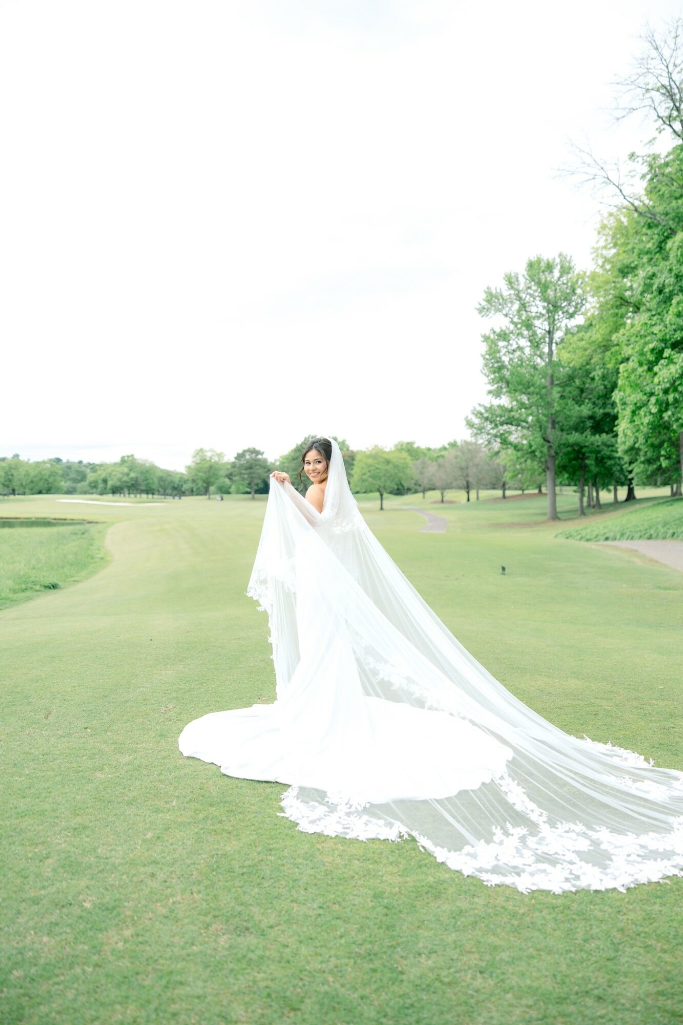 Woman in a wedding dress holding a long veil outdoors on a grassy field with trees, under a bright sky.