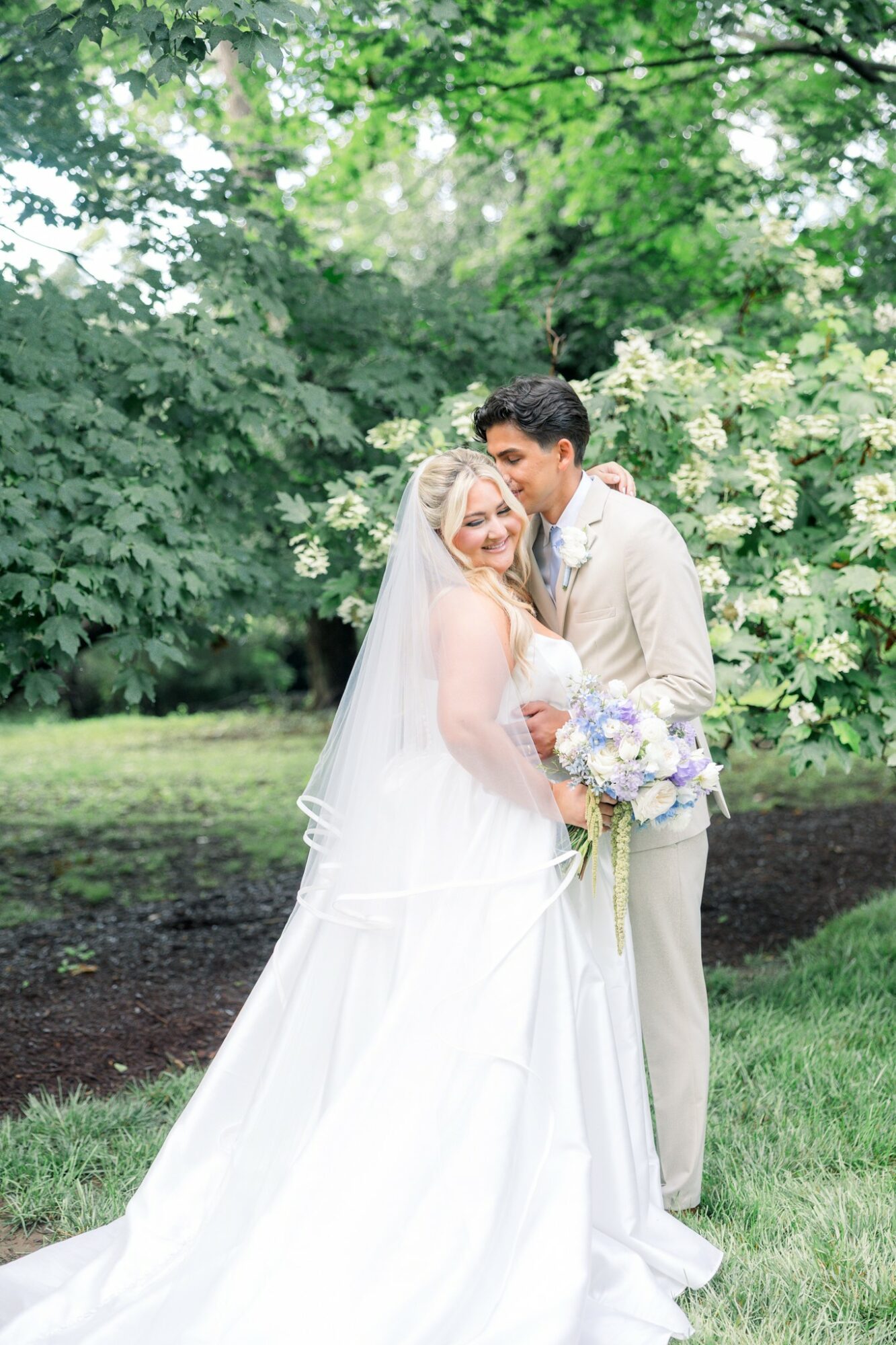 A bride and groom embrace outdoors among green trees and white flowers, smiling at each other, bride holding a bouquet.