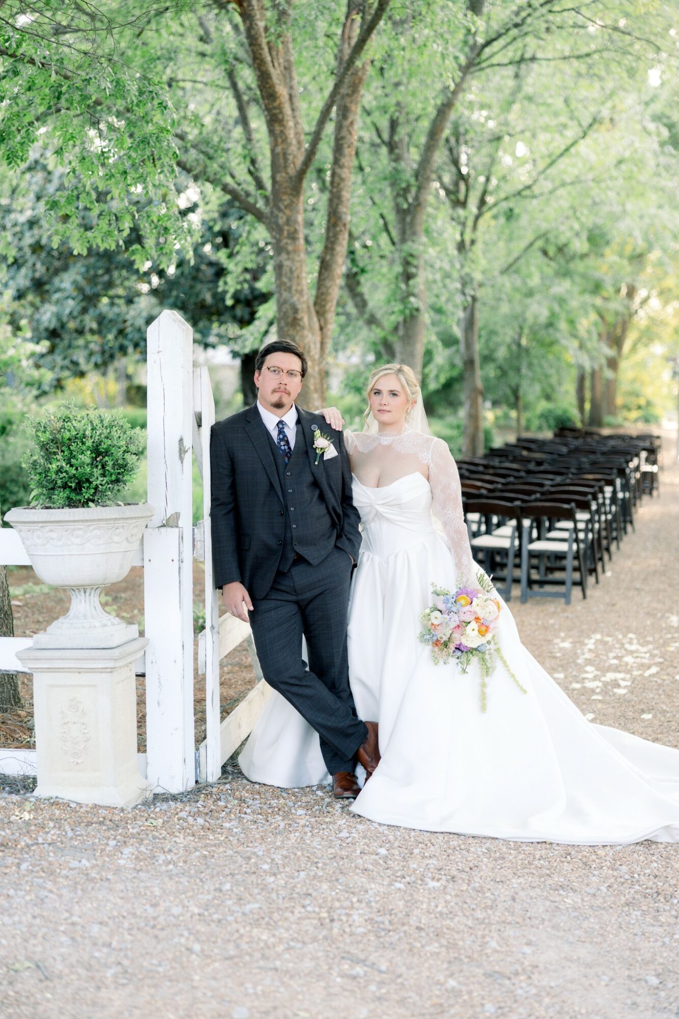 Bride and groom standing outdoors near a white gate, trees in background, bride holding a bouquet, groom in suit.