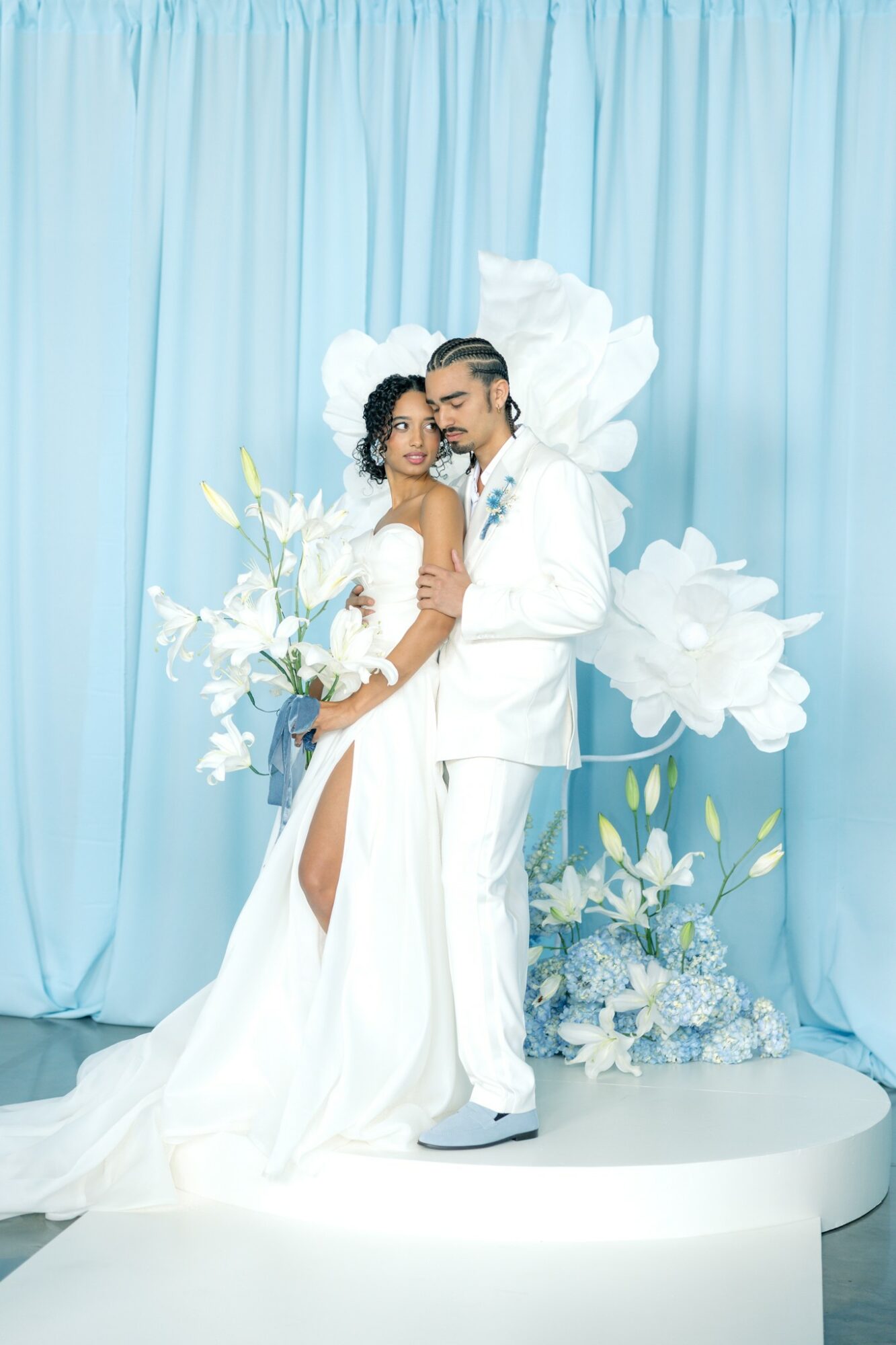 Bride and groom in wedding attire stand close together with floral backdrop, light blue curtain, and white platform behind them.