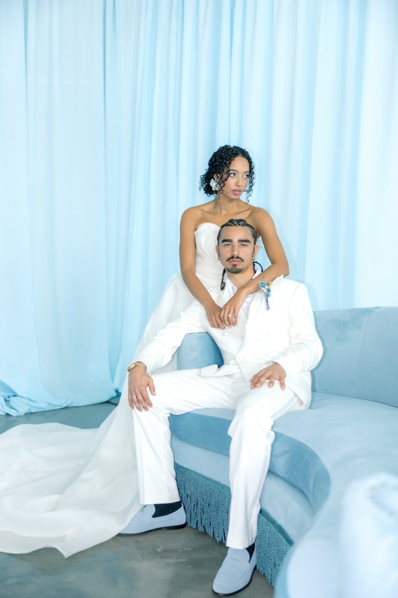 A woman in a white dress stands behind a seated man in a white suit on a sofa, with light blue curtains in the background.