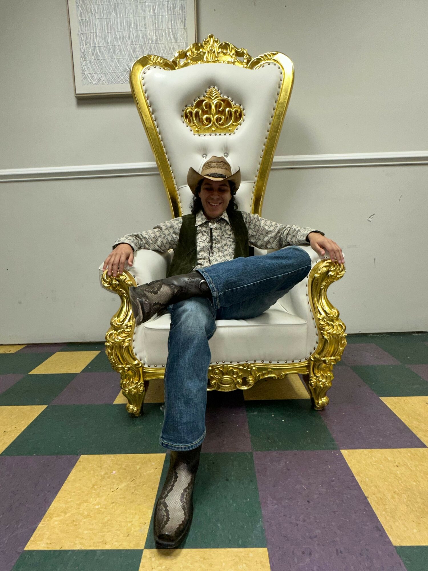 Person sitting on a white and gold ornate throne with a high back, in a room with colorful checkered floor.