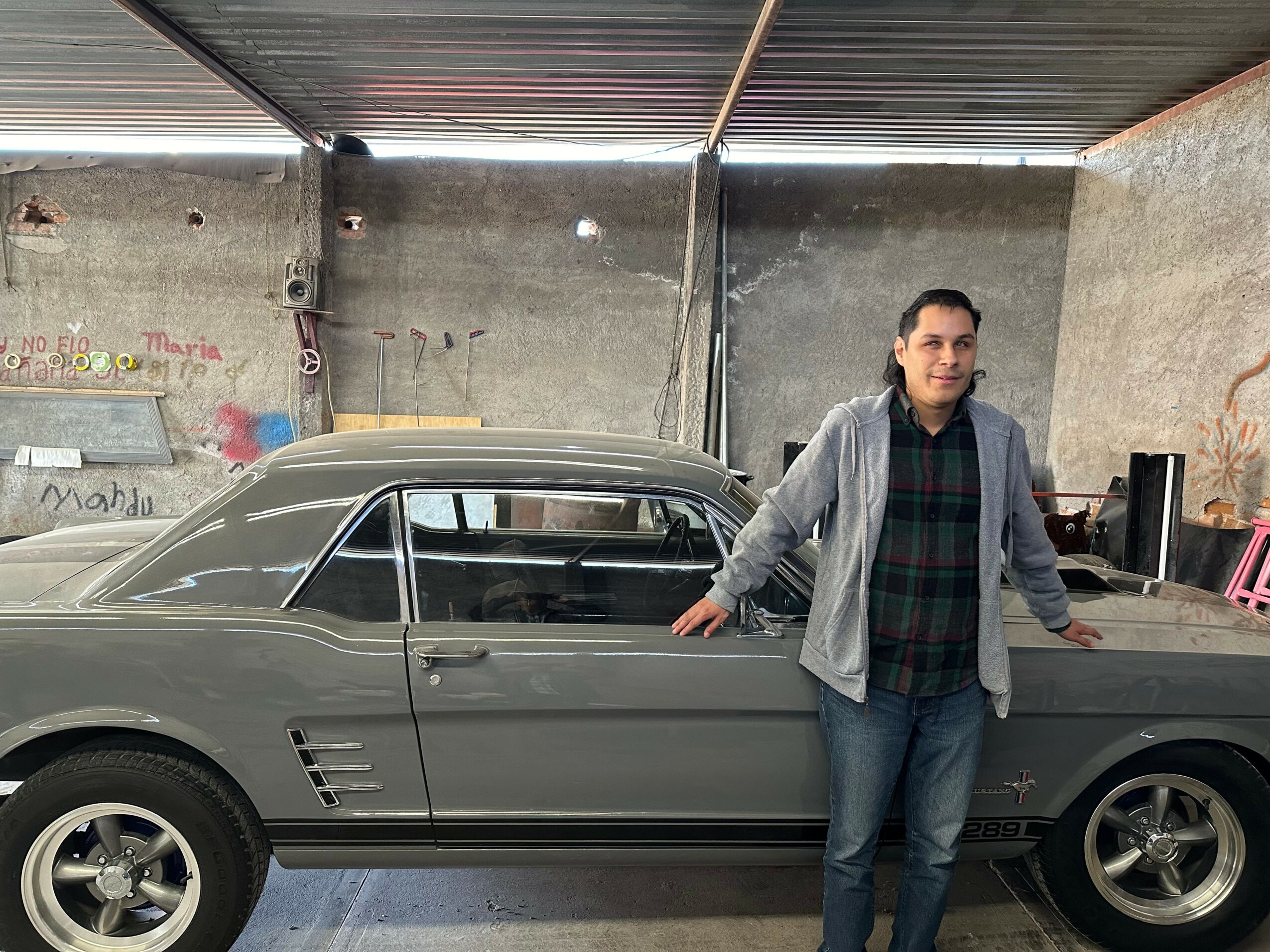Man standing next to a classic gray car inside a garage, with a concrete wall and metal roof.
