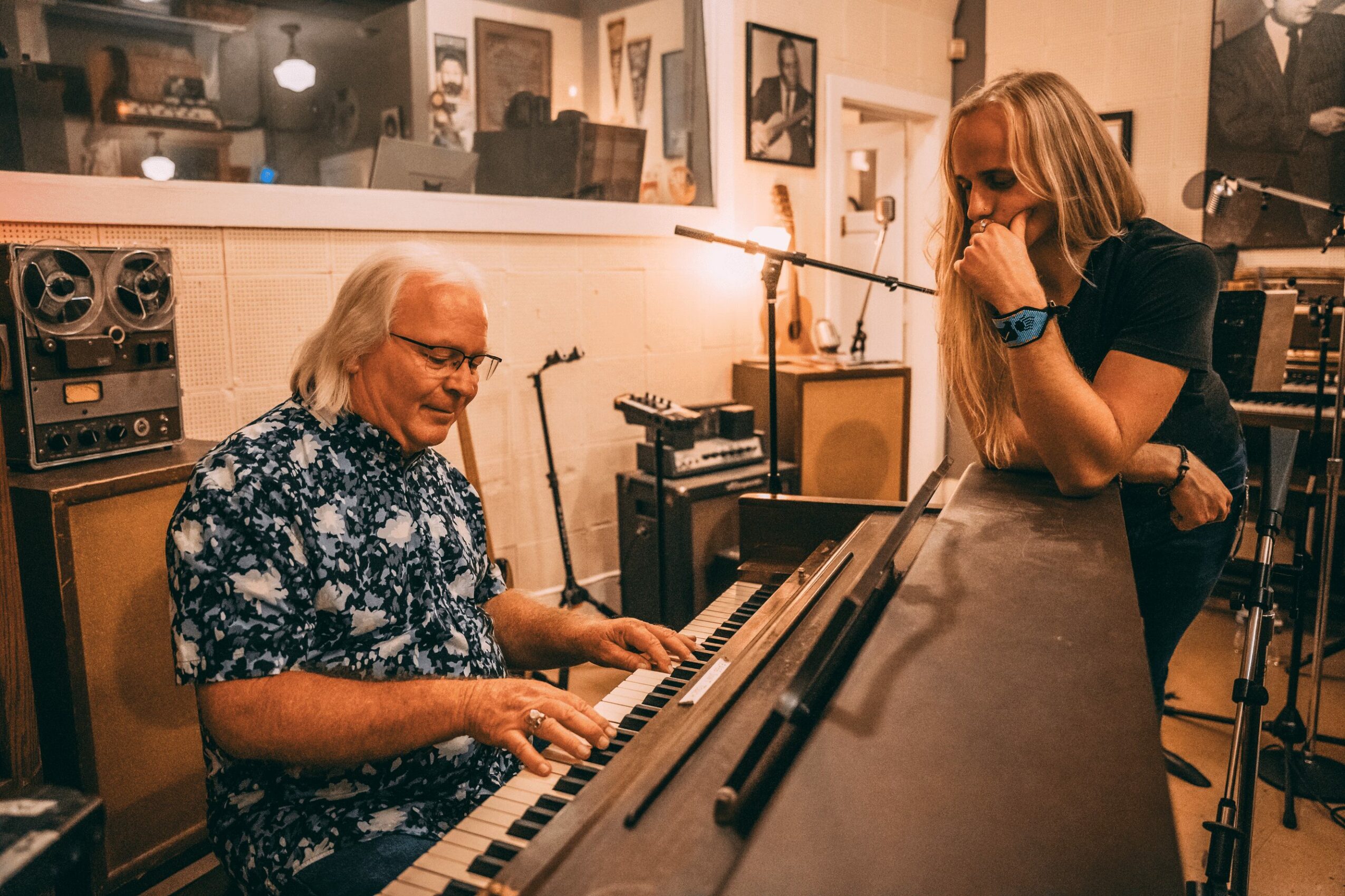 A man playing piano with a woman watching, in a room with framed photos on the wall.