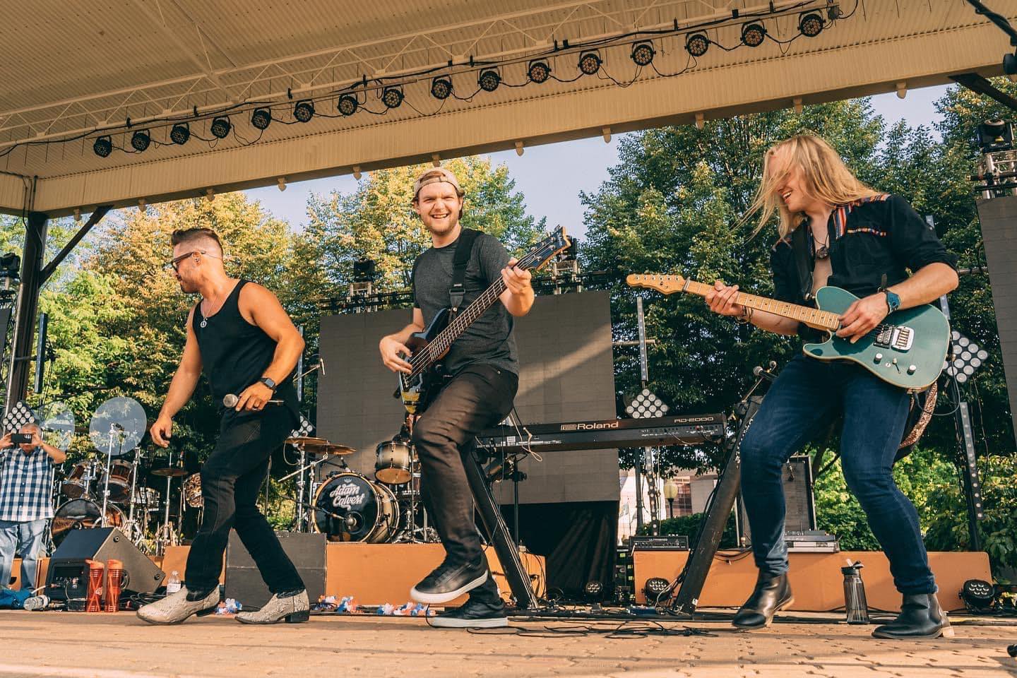 Three musicians perform on an outdoor stage with trees in the background, playing instruments and singing.