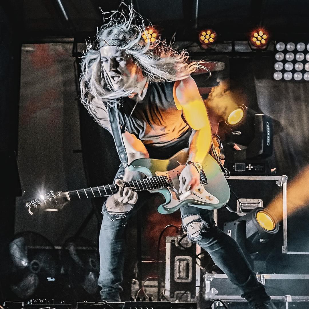 Musician with long hair playing an electric guitar on stage, with stage lights and equipment in background.