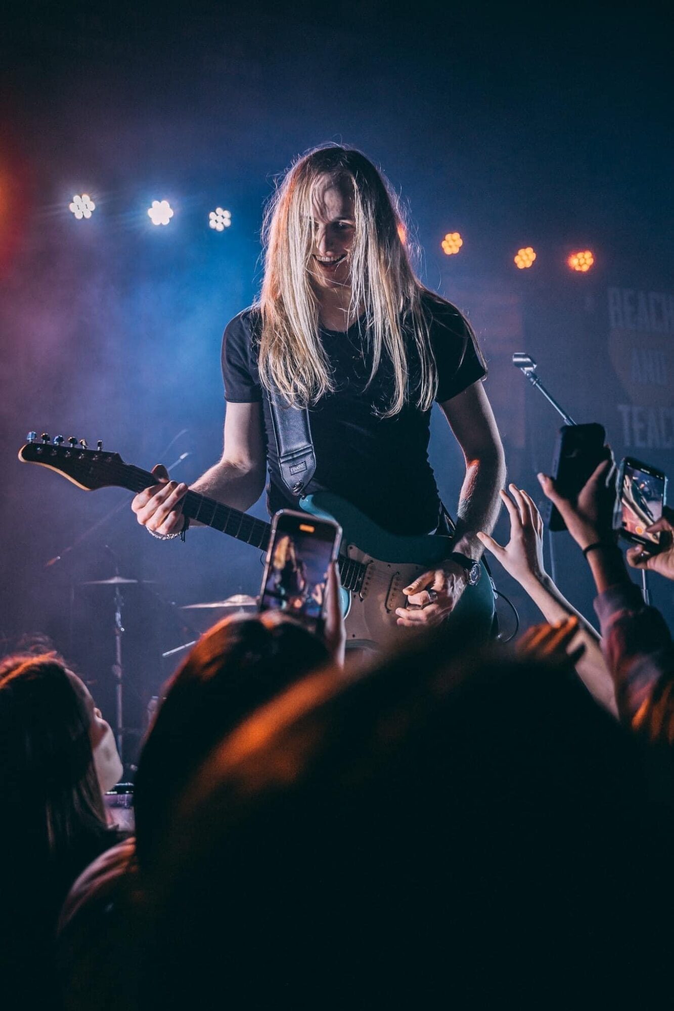 Female musician with long hair playing guitar on stage with audience taking photos, stage lights in background.