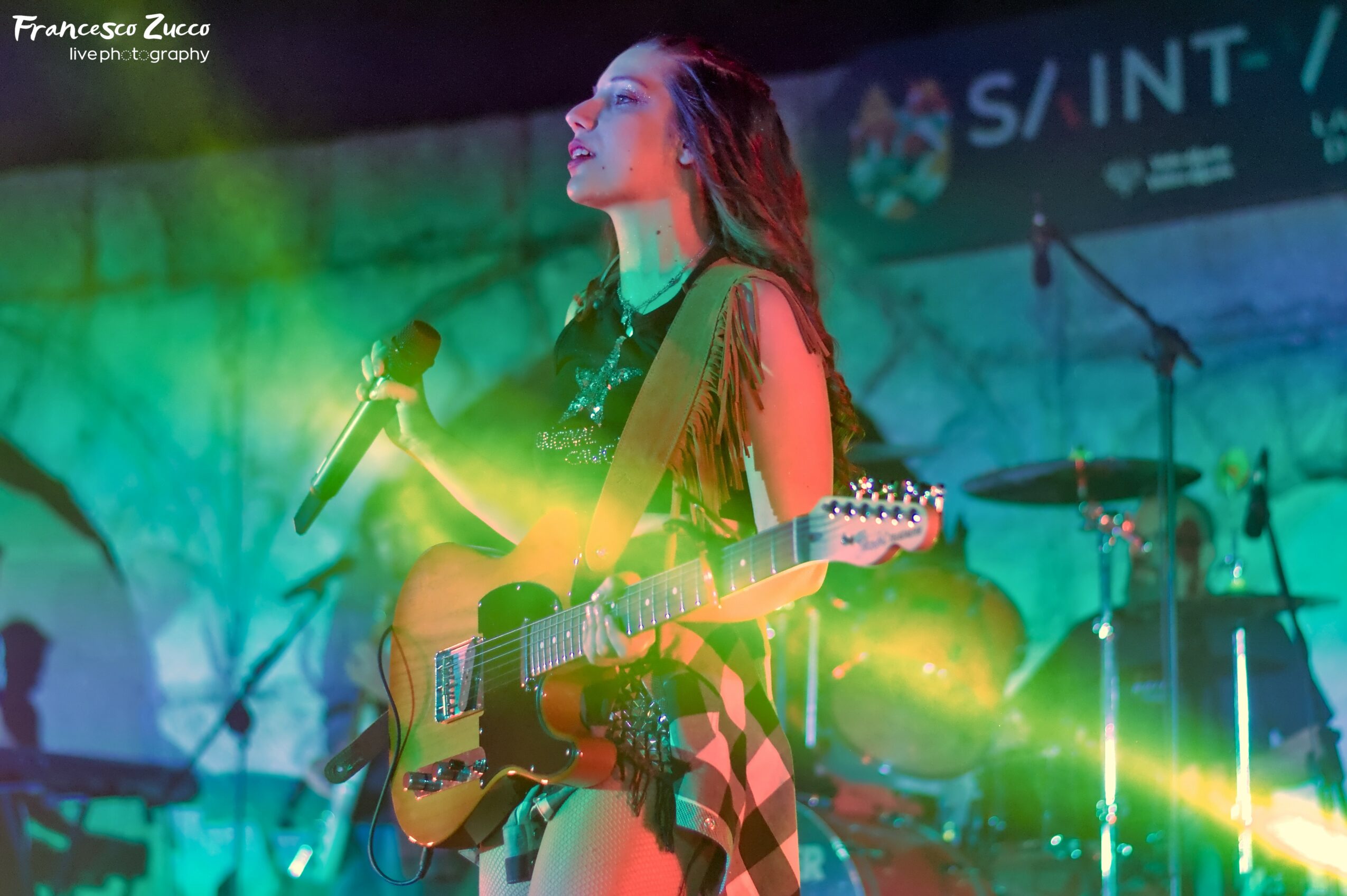 Female musician with long hair holding a guitar and microphone on stage with colorful lighting.
