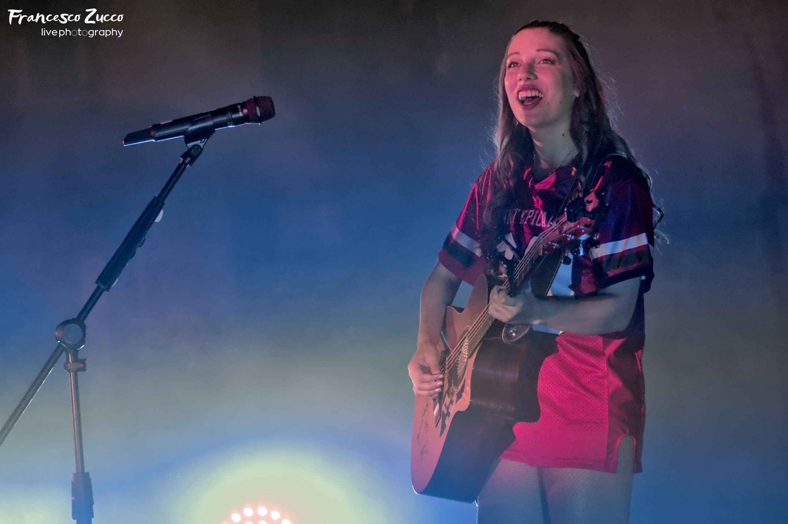 Young woman with long hair singing and playing guitar on stage with a microphone, smiling, with colorful stage lighting.