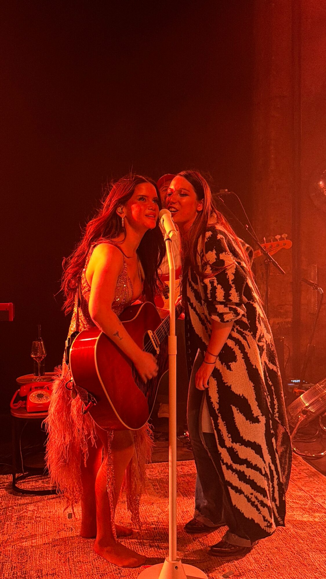 Two women singing into a microphone on stage, one playing guitar, with red lighting and musical equipment nearby.