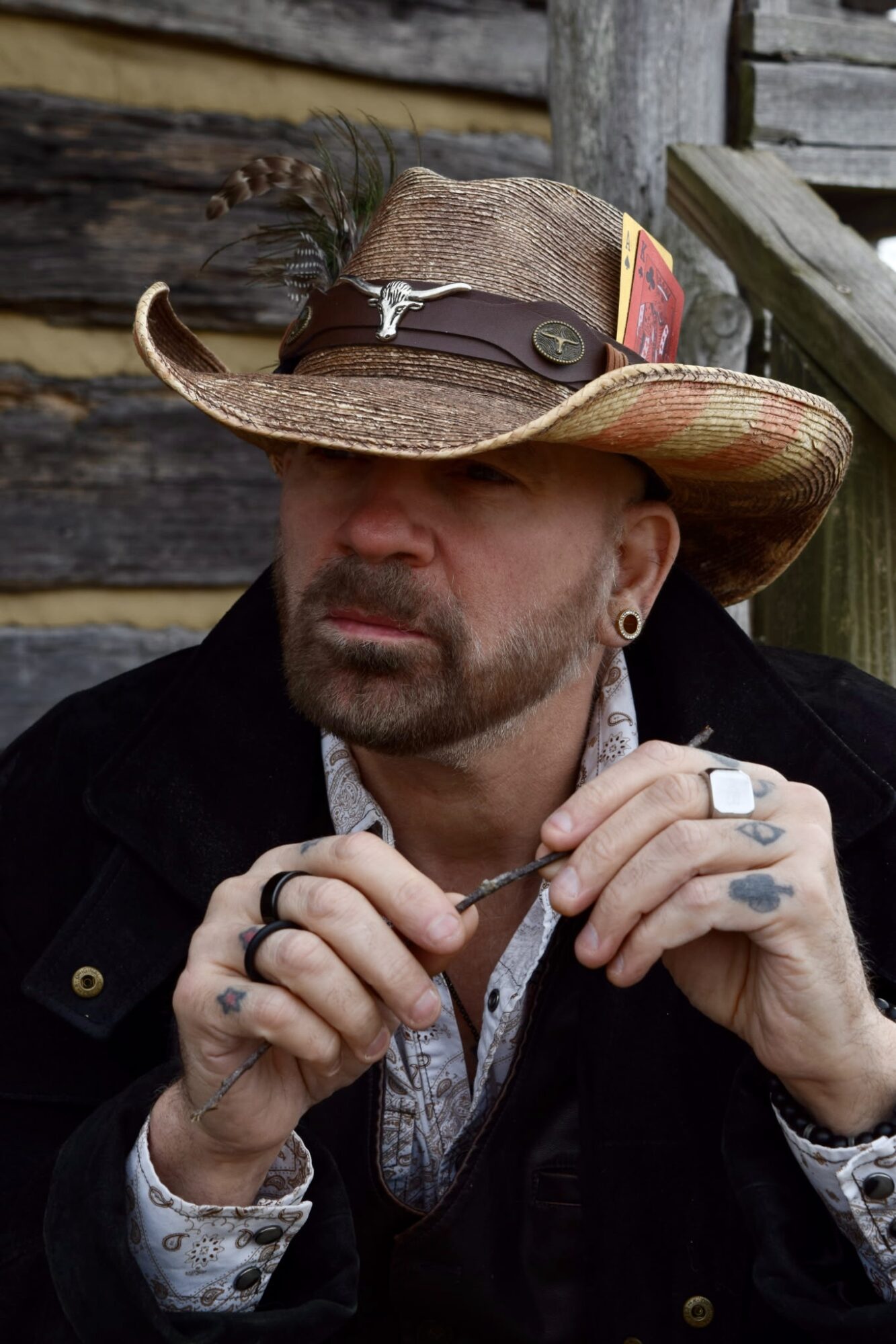 Man wearing a cowboy hat with a feather, sitting outdoors with a wooden background, holding a cigarette, with a serious expression.