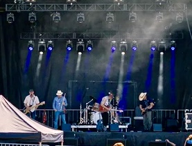 Band performing on stage with five musicians, stage lights, and fog effects, audience visible at the bottom.