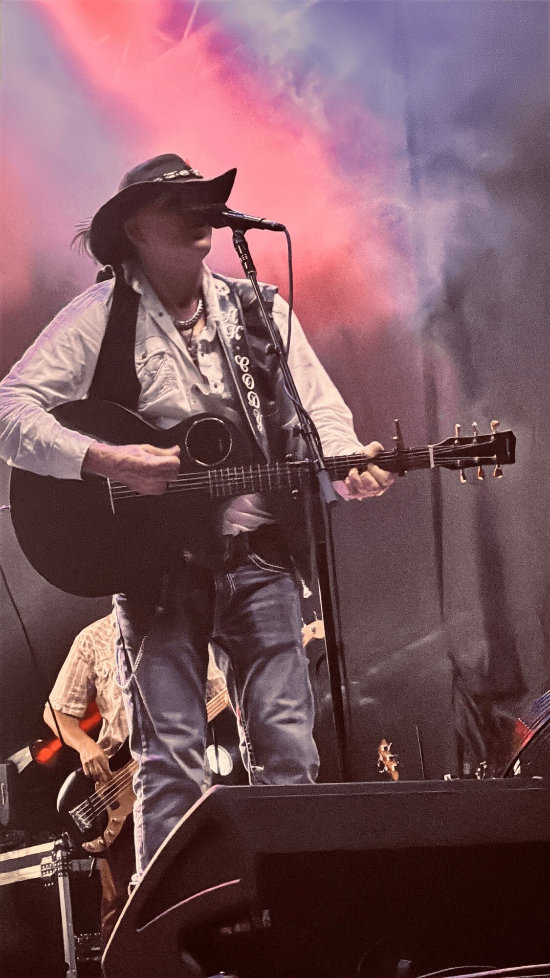 Person wearing a hat and sunglasses playing guitar and singing into a microphone on stage with colorful smoke in background.