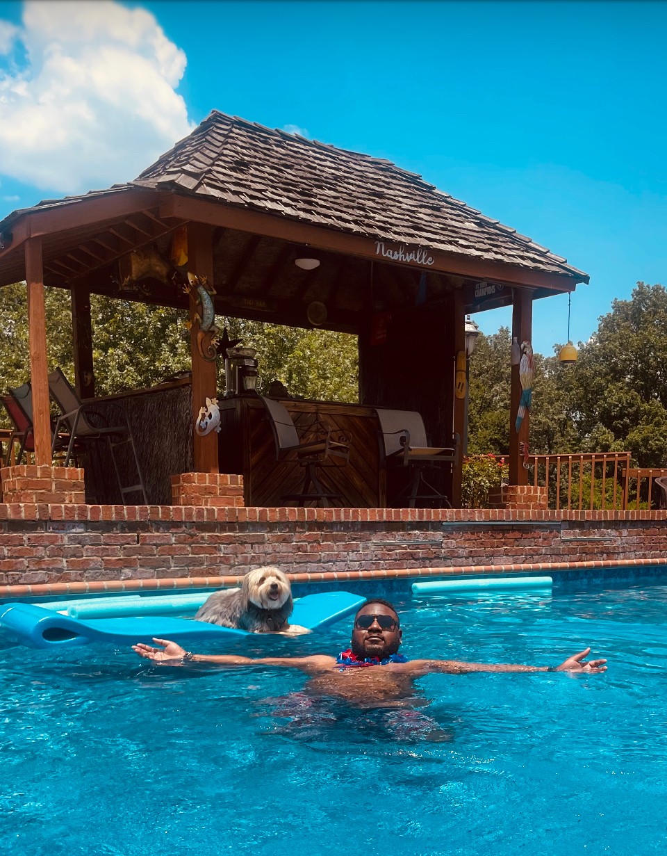 Two people in a swimming pool, one with a dog, with a wooden pavilion and trees in the background.