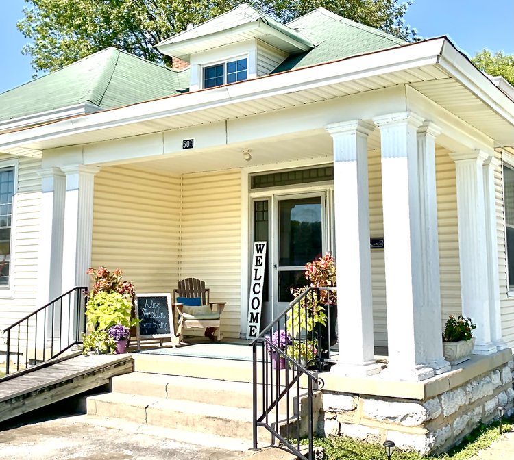 Front porch of a house with a welcoming door, two chairs, potted plants, and a welcome sign, under a green roof.