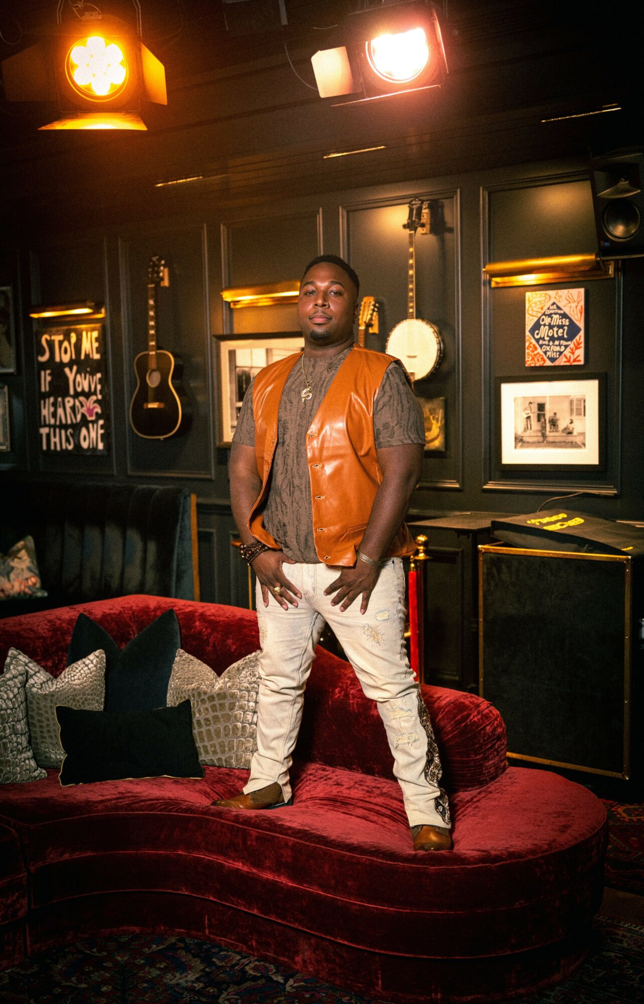 Man standing on a red velvet sofa in a dimly lit room with framed pictures and musical instruments on the wall