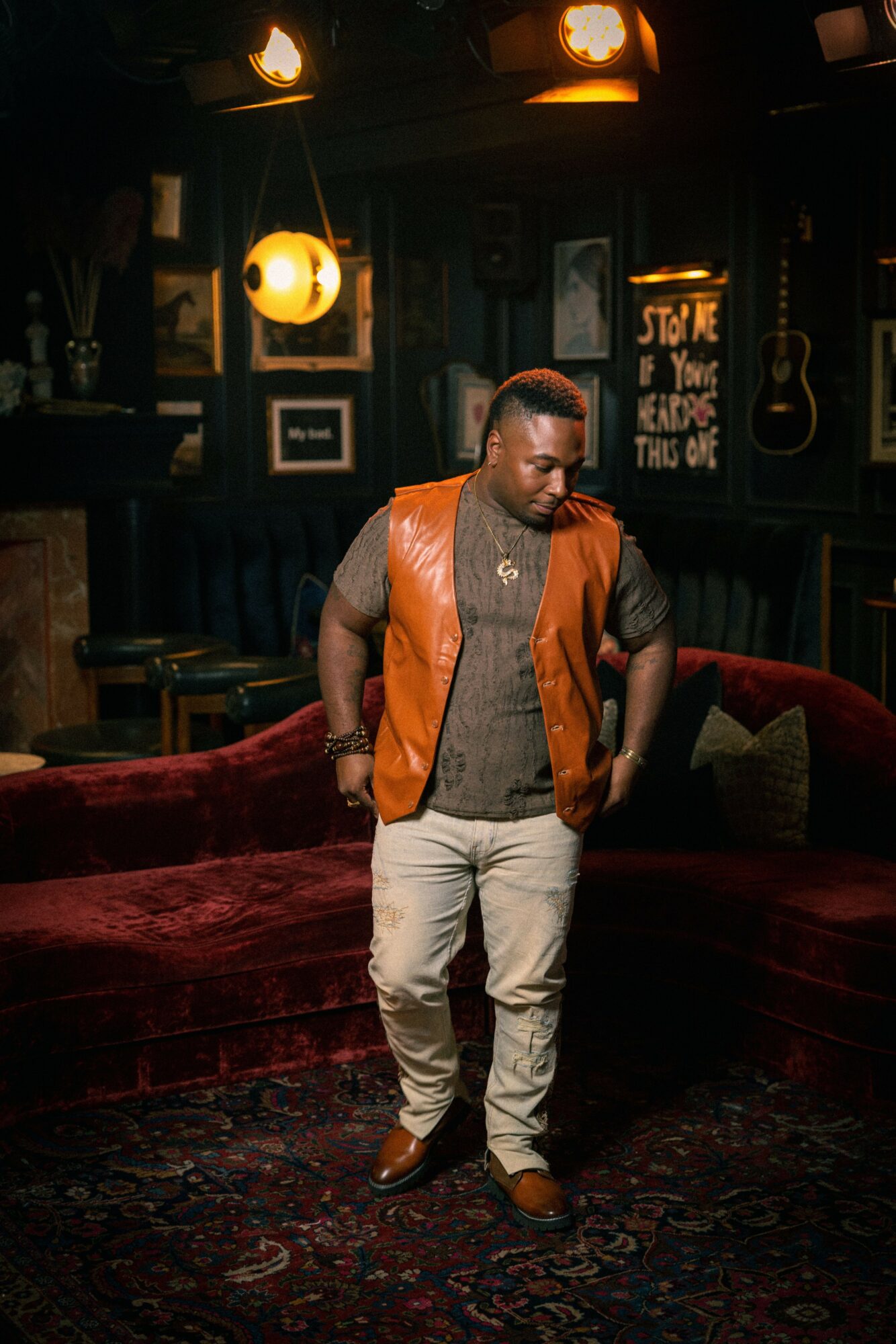 Man standing in a dimly lit room with dark walls, framed pictures, and a chalkboard sign, wearing a brown vest and light pants.