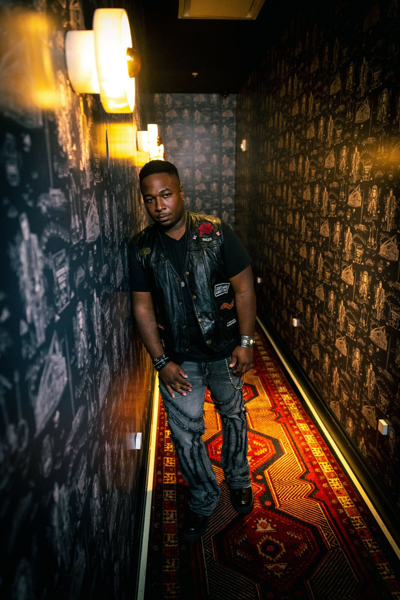 Man standing in a narrow hallway with dark patterned walls and a red patterned carpet, illuminated by wall lights.
