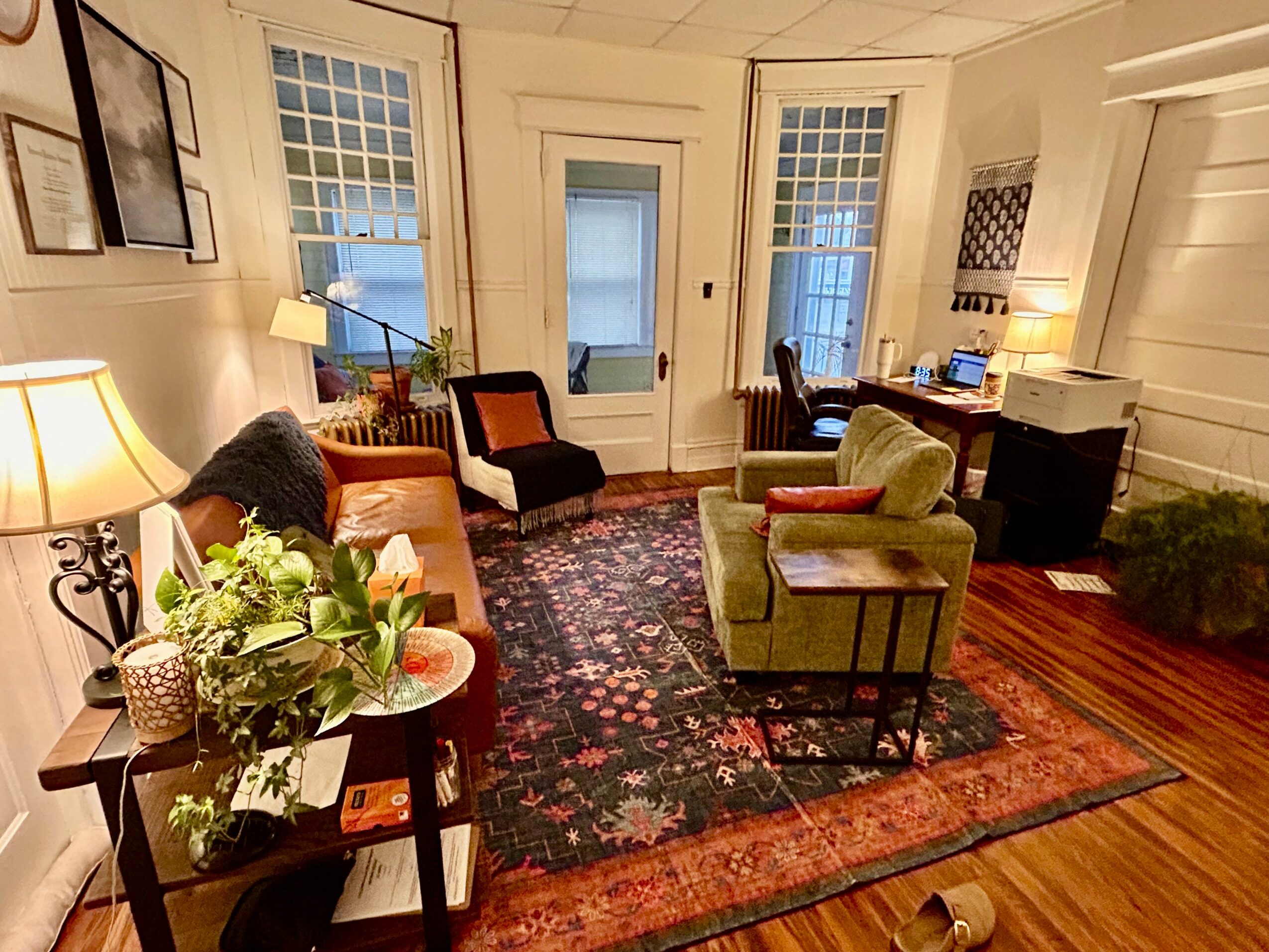Living room with chairs, a desk, and a floral arrangement on a table, illuminated by lamps, with windows and hardwood floors.