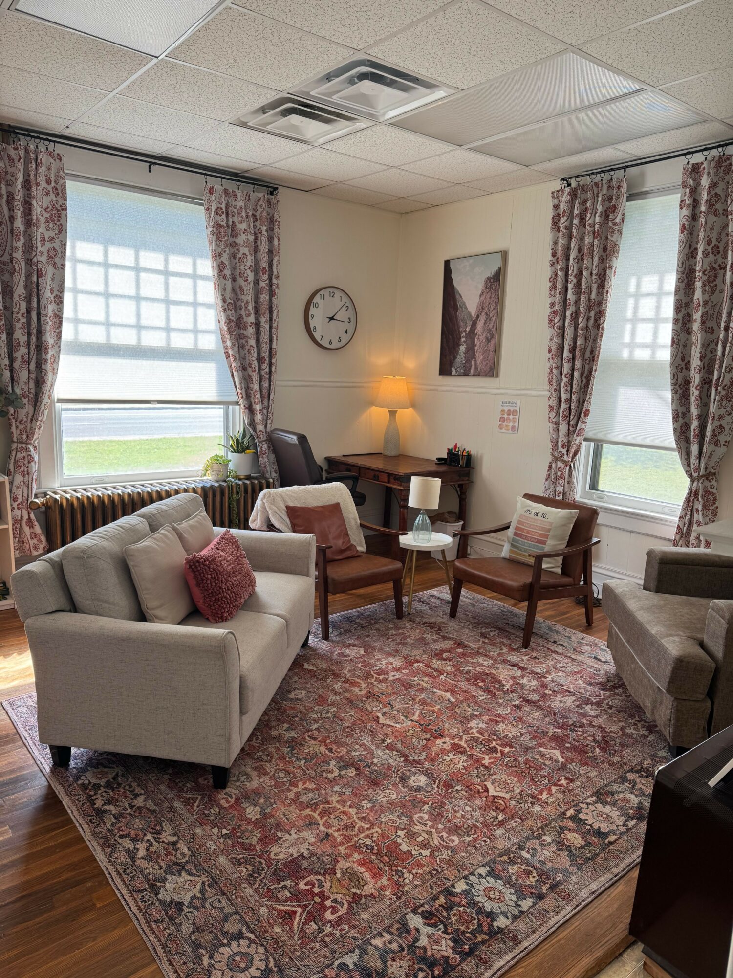 Living room with a white sofa, armchair, wooden table, and large window with curtains, sunlight, and a clock on the wall.