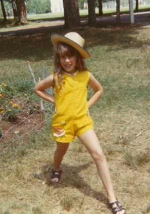 Young girl in yellow outfit and wide-brimmed hat standing outdoors with hands on hips, smiling, in a grassy area with trees.