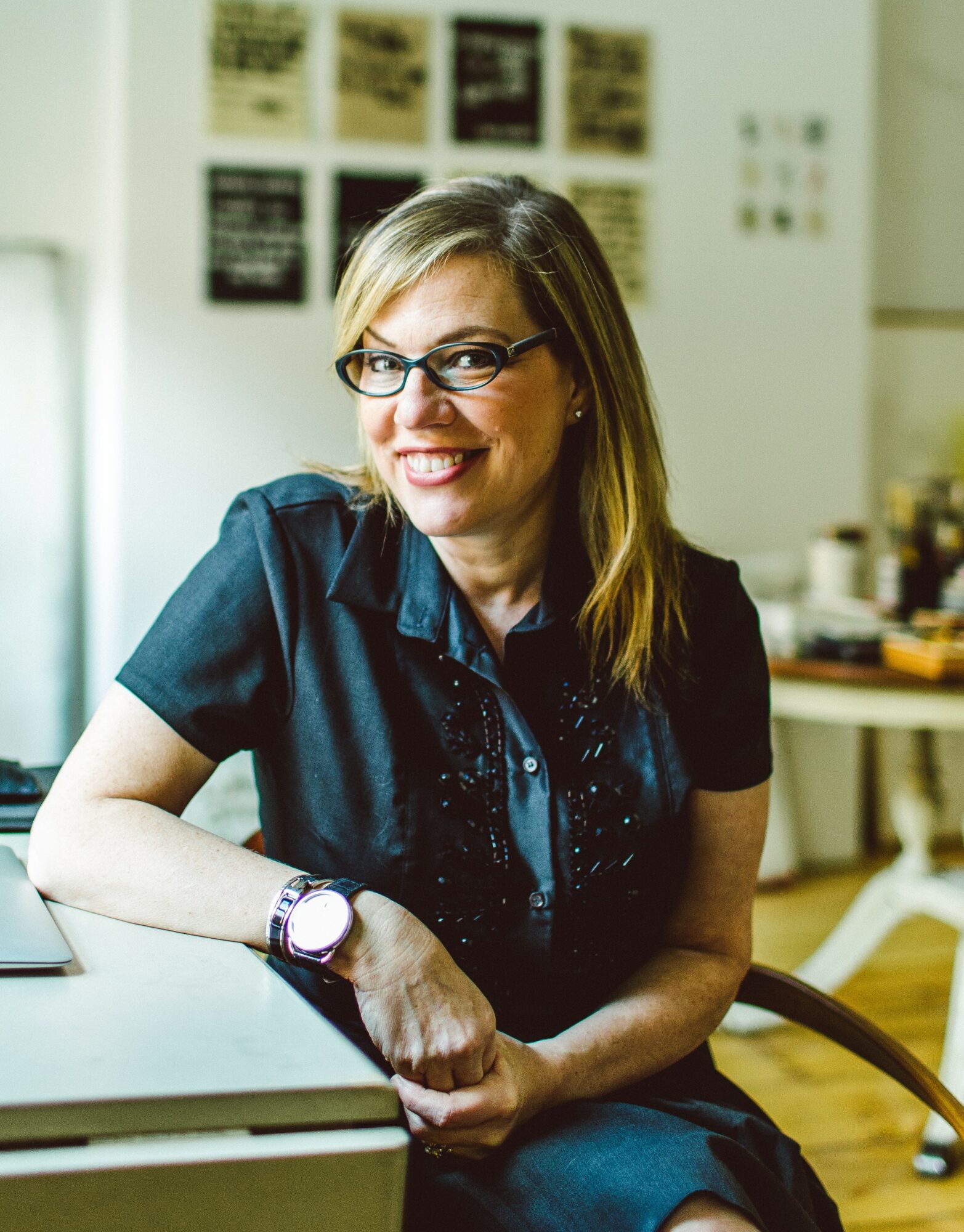 Woman with glasses smiling, sitting at a desk in a room with posters on the wall, wearing a dark shirt.