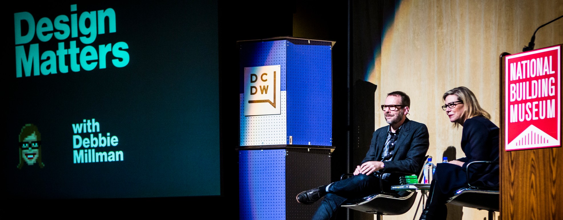 Two people seated on stage with a screen displaying 'Design Matters' and logos, in a presentation setting.